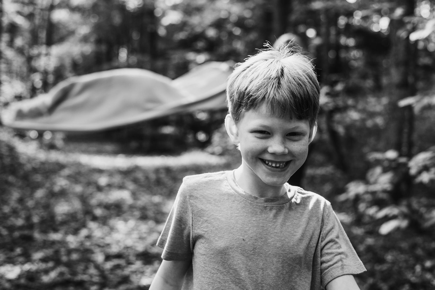 Black and white portrait of a joyful child outdoors wearing a light-colored t-shirt, with foliage in the background.