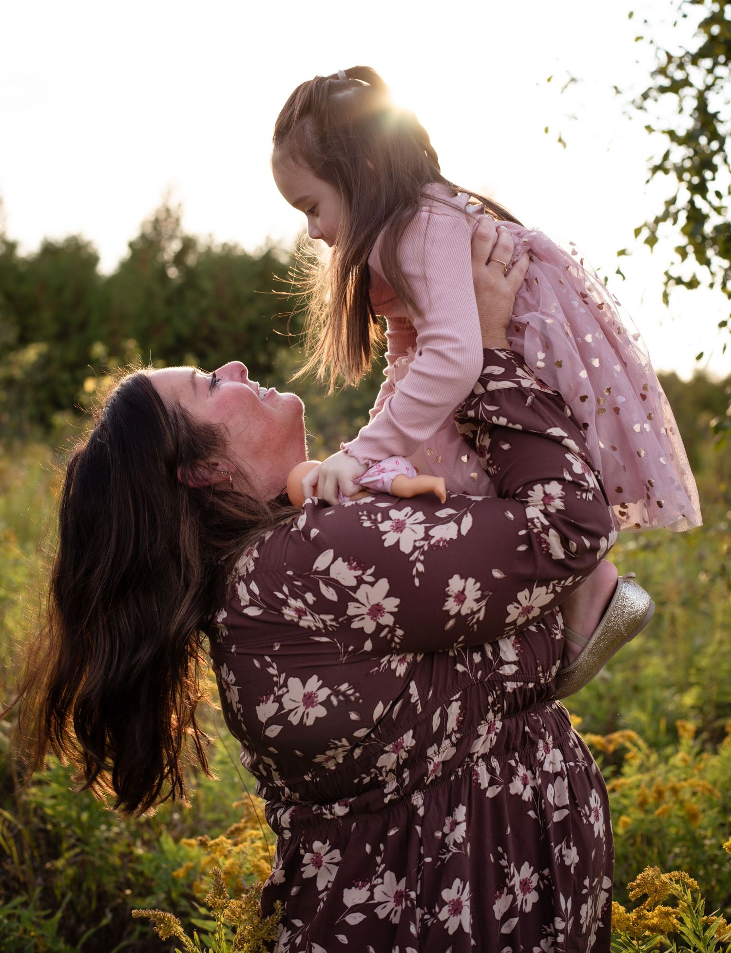 A mother lifts her young daughter in a sunny field, both smiling, wearing floral and pink outfits.