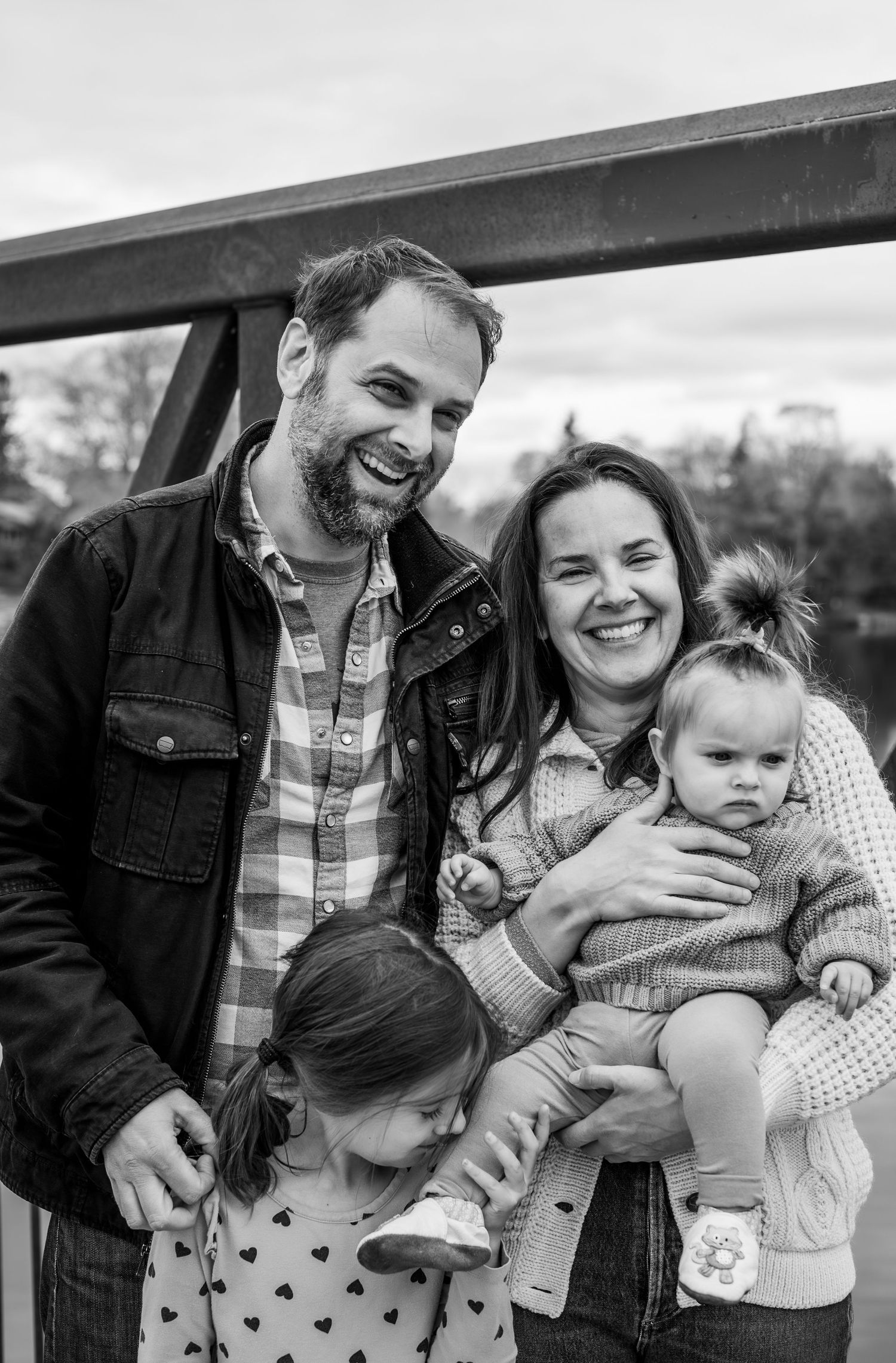 Black and white family portrait of smiling couple with two young daughters on a bridge outdoors.