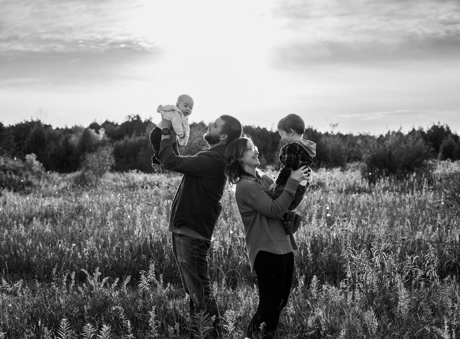 Black and white photo of a couple lifting two babies in a field at sunset.