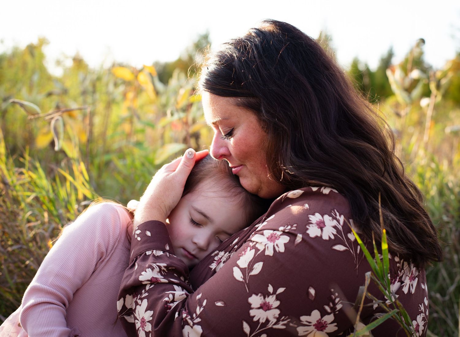 A mother tenderly embraces her young daughter in a sunlit autumn field, sharing a peaceful moment.