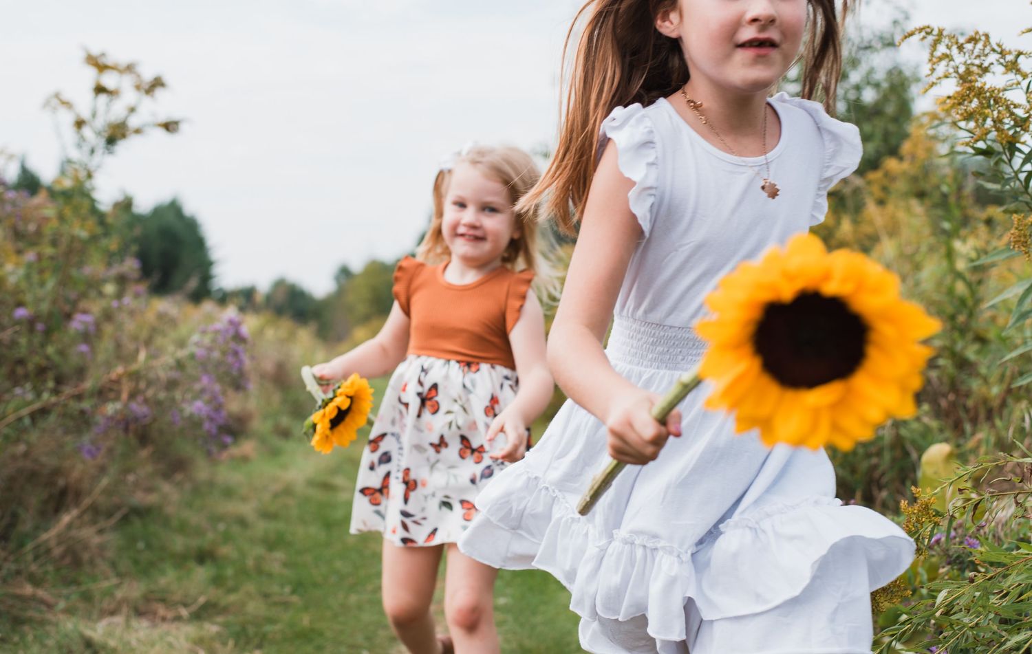 Two young girls running through a field holding bright sunflowers on a sunny day.