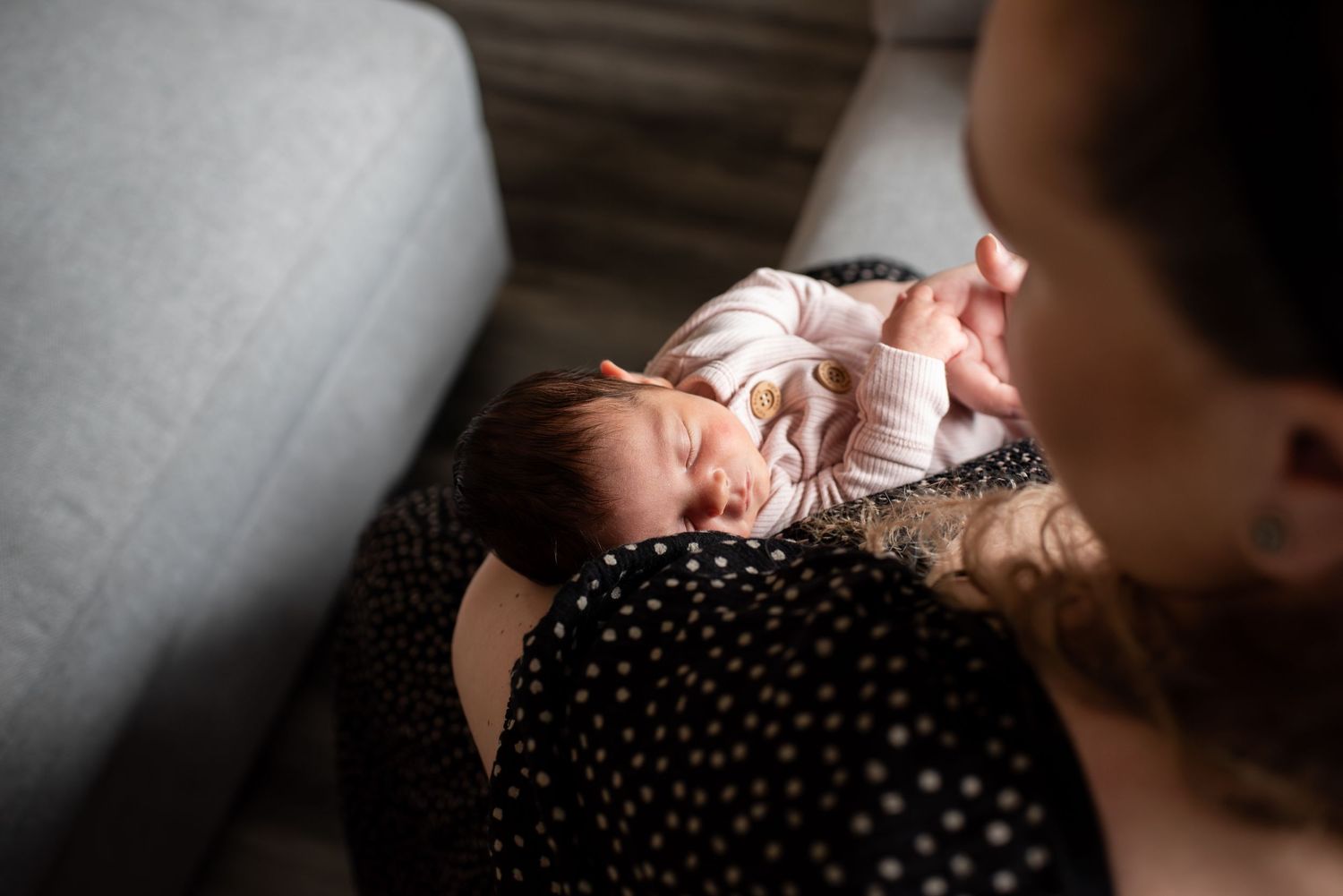 Parent snuggling with a newborn baby on a gray couch in gentle natural light.