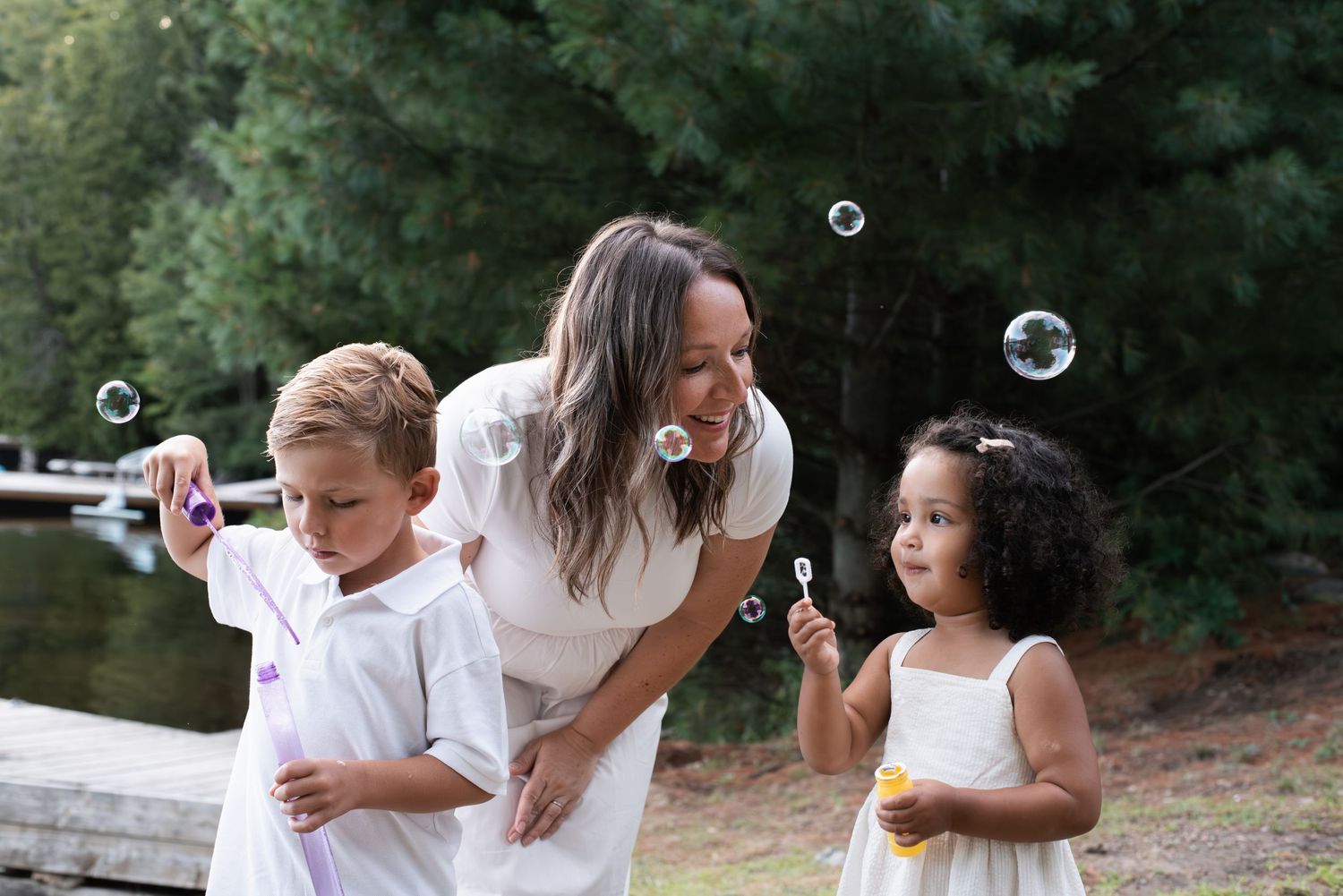 A woman and two children blowing bubbles outdoors near a lake surrounded by trees.