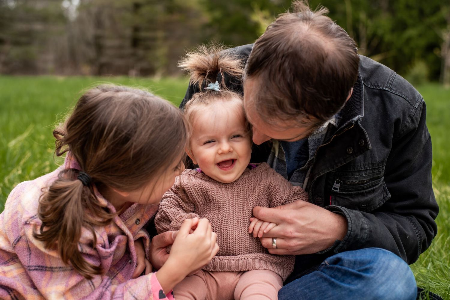 A joyful baby smiles as parents lean in for kisses outdoors on green grass in a heartwarming family moment.