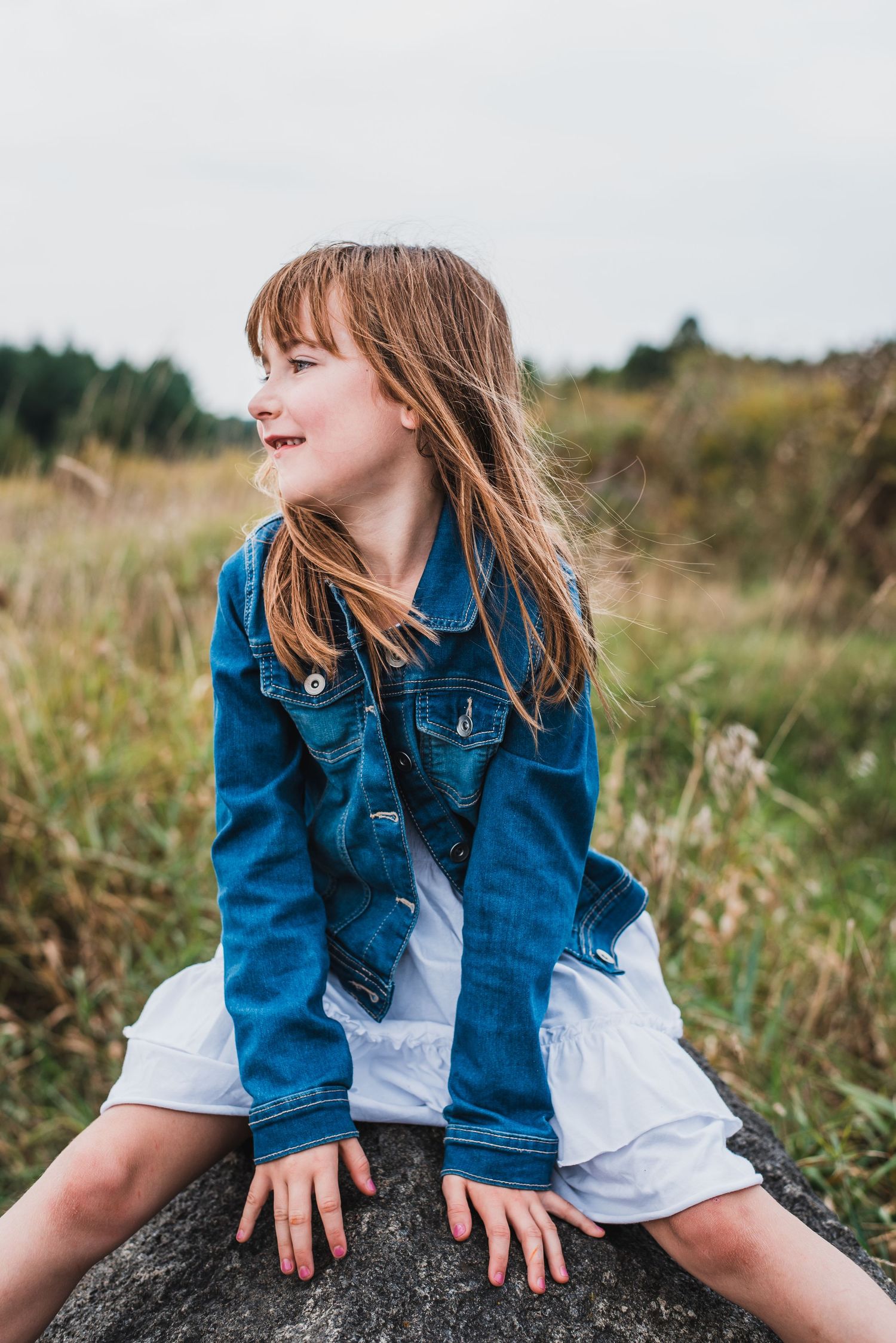 In a grassy field, a young child sits atop a rock wearing a denim jacket and white dress while smiling.