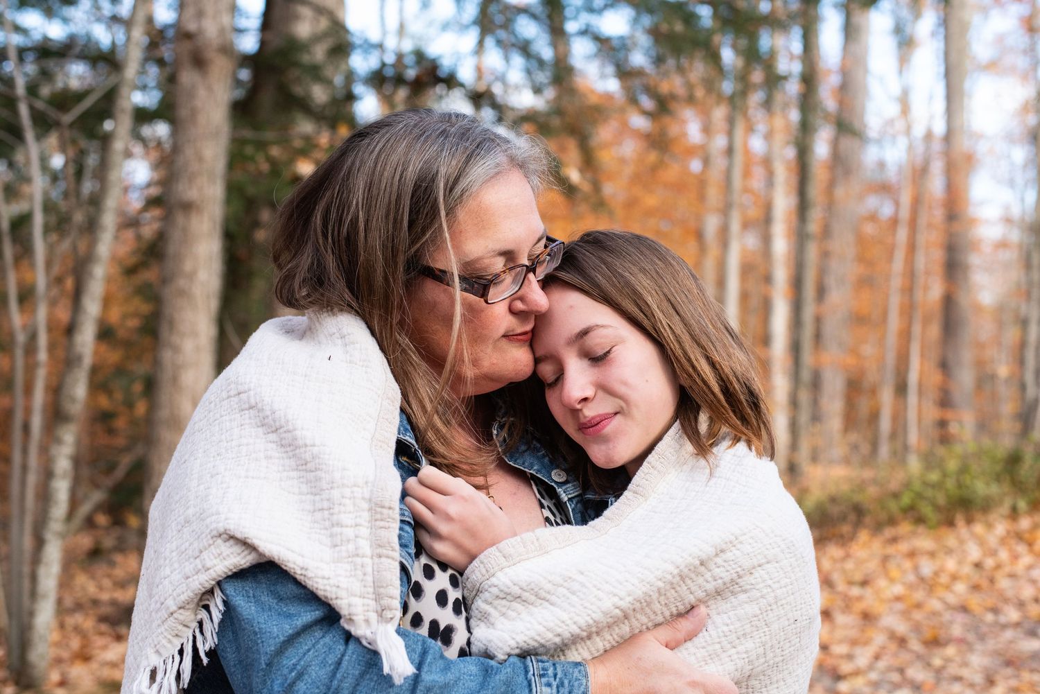Two people share tender moments in a wooded forest area during autumn with fallen leaves all around them.
