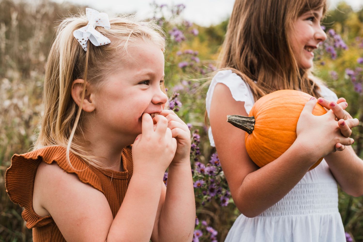 Two young girls holding orange pumpkins among purple wildflowers in an autumn field.