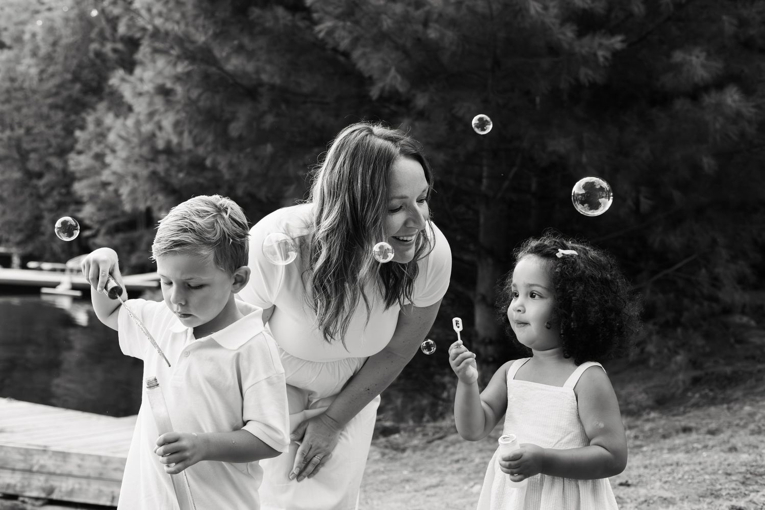 Black and white photo of a woman and two children blowing bubbles outdoors near a lake.