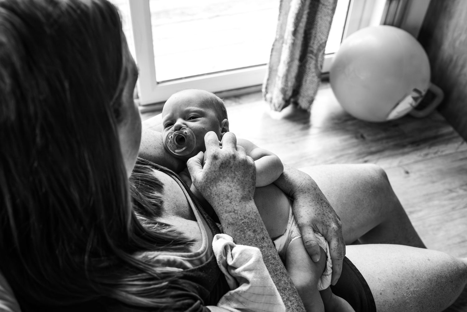 Black and white photograph of a baby with a pacifier being cradled on a sofa near a window.