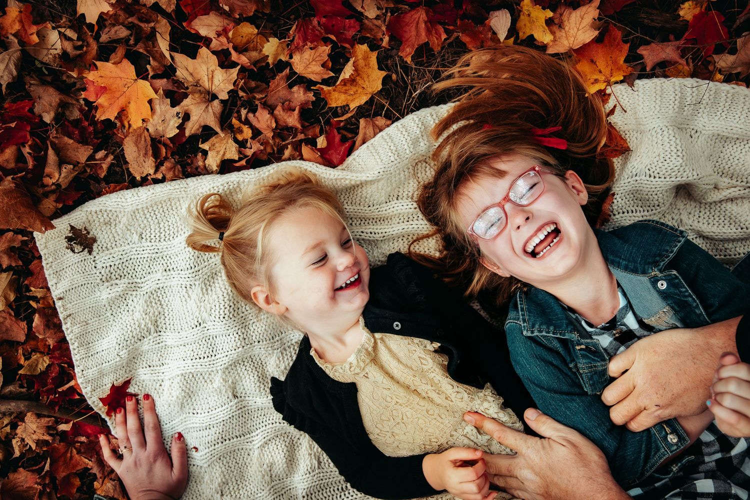 Two joyful children laughing while lying on a blanket surrounded by colorful autumn leaves outdoors.