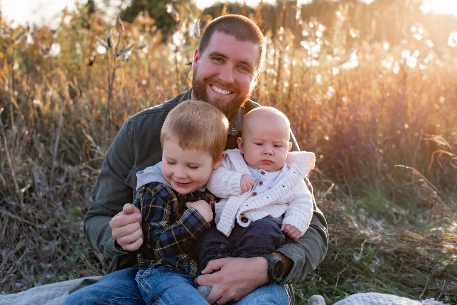 A smiling bearded man sits outdoors holding two young children, a toddler and an infant, in a sunlit field.