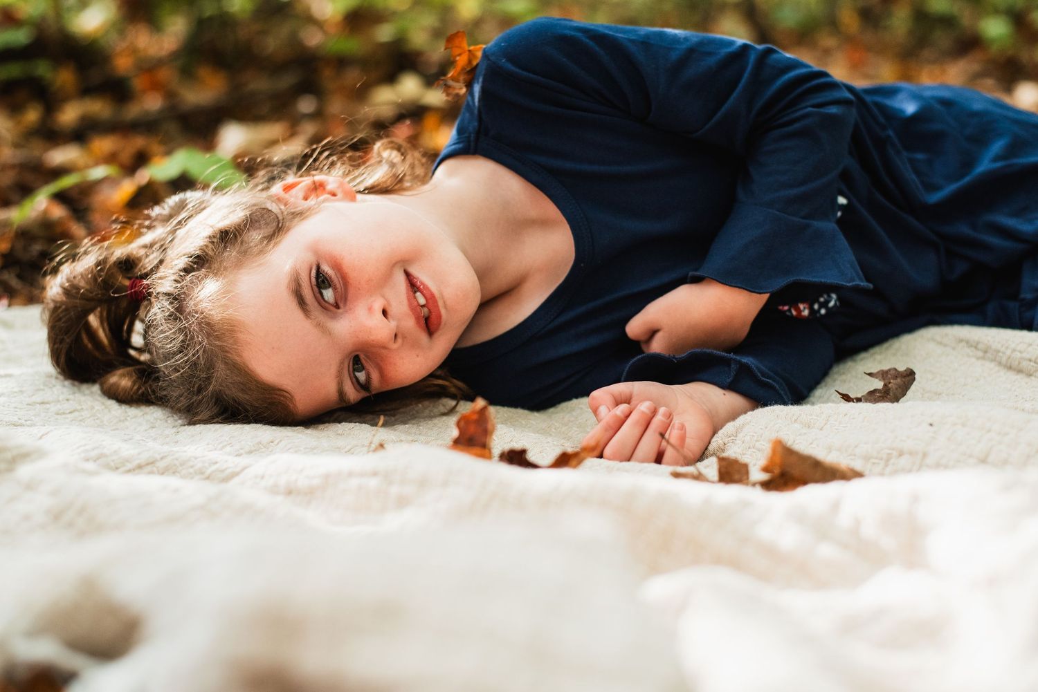 A figure lies on a white fabric surface surrounded by scattered autumn leaves and natural outdoor lighting.