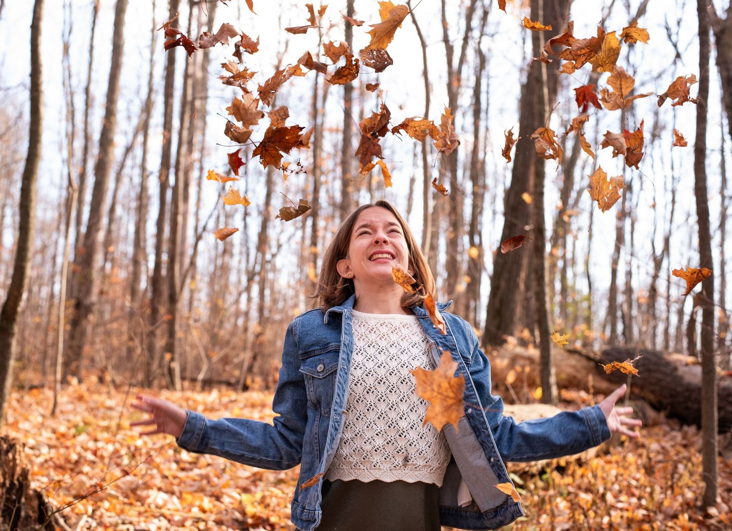Person joyfully throws autumn leaves in the air while sitting on ground in a forest during fall season.