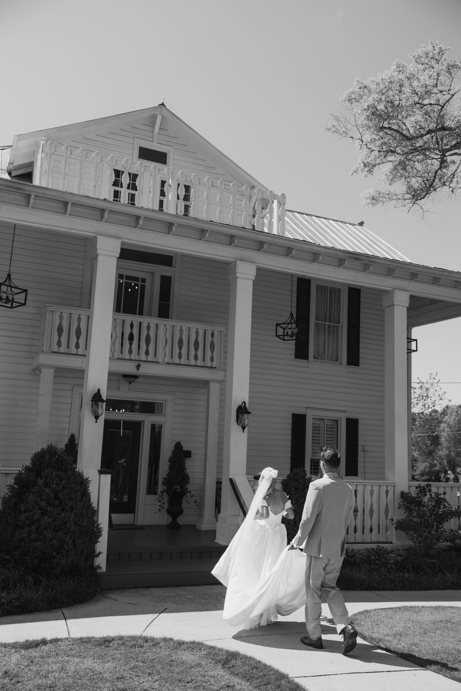 Classic white colonial mansion with wrap-around porch and elegant decorative details serves as backdrop for a wedding celebration.