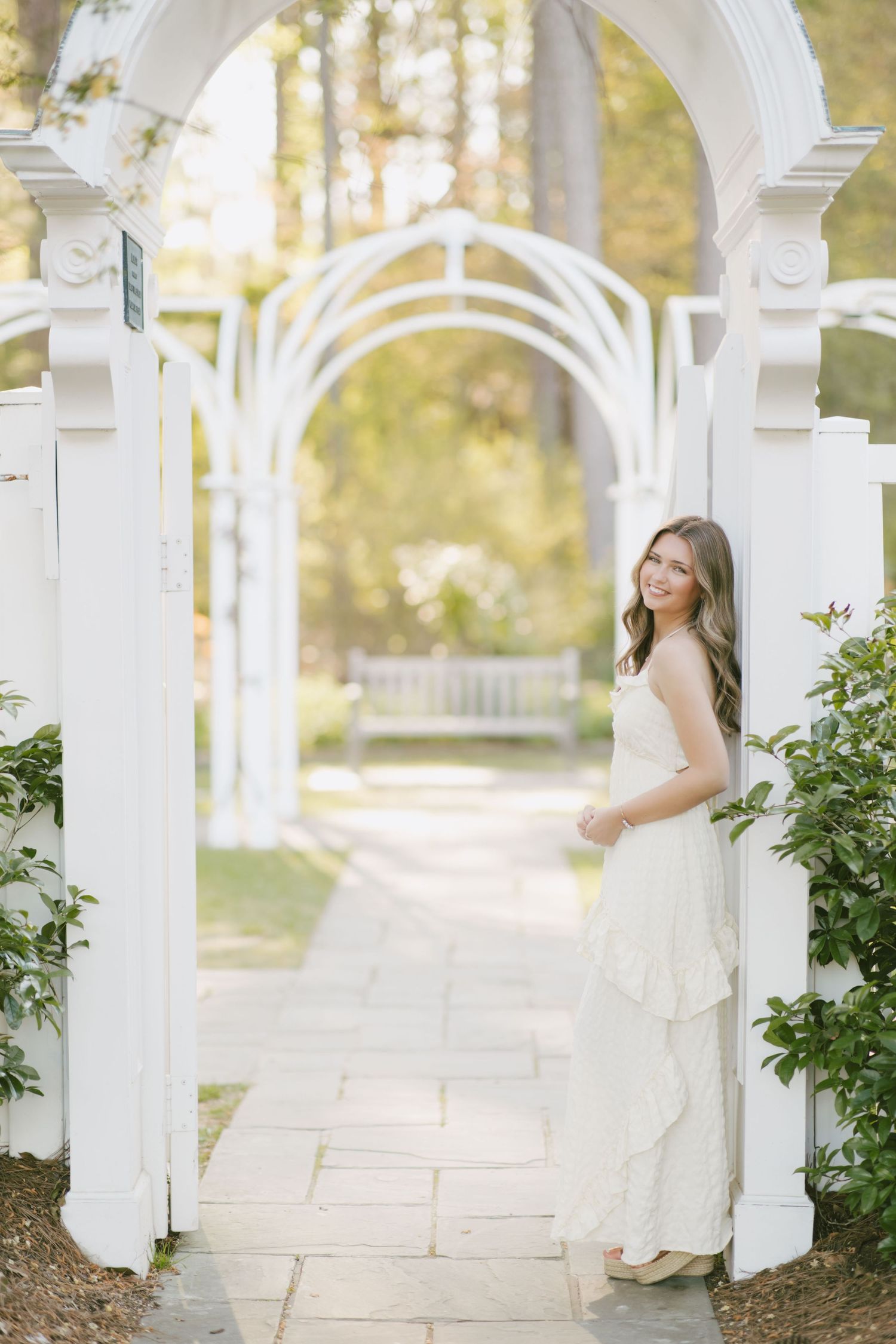 A white dress flows elegantly under ornate garden archways with stone paths and lush greenery in soft sunlight.
