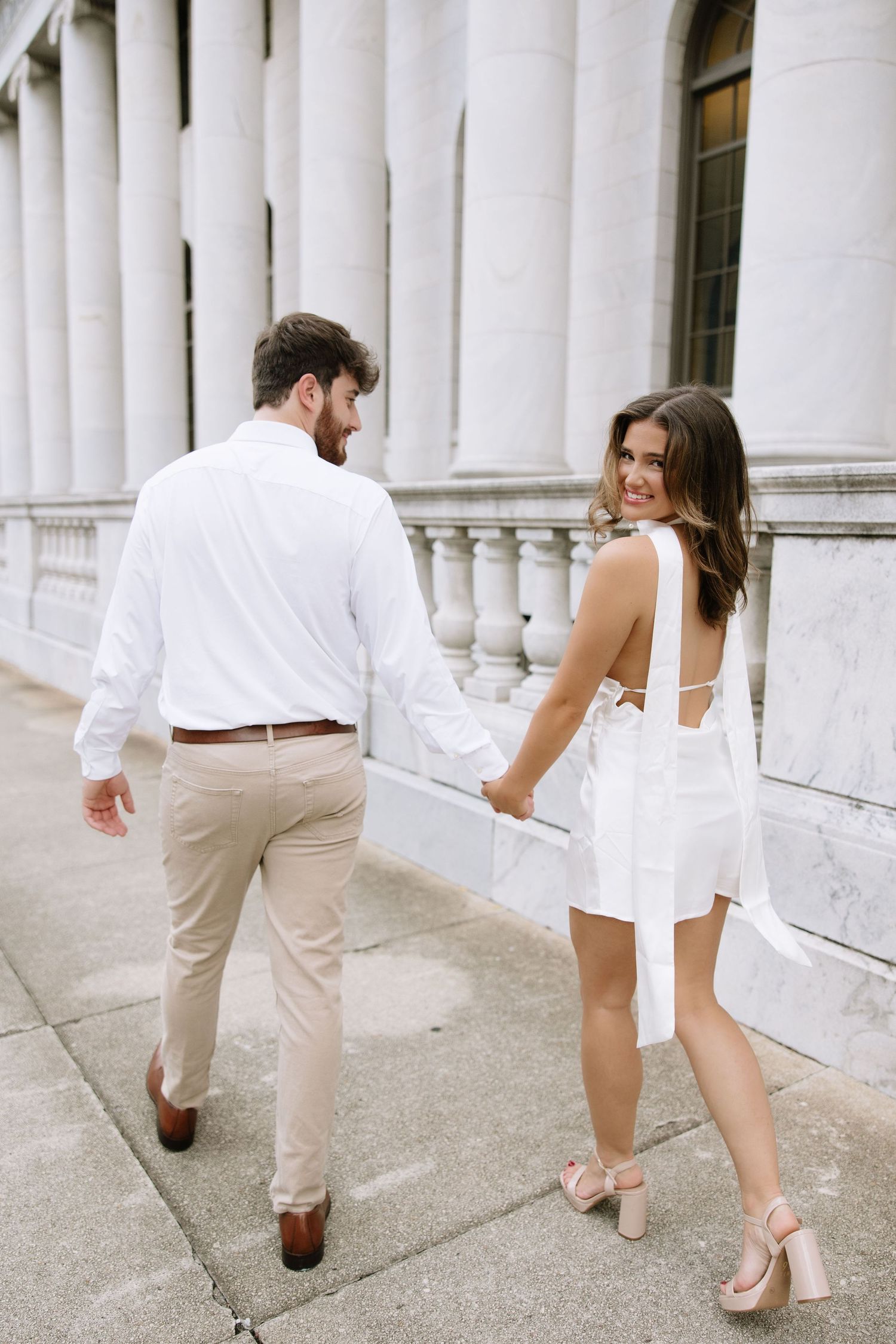A couple in coordinated white and beige outfits walk together along a columned building exterior.