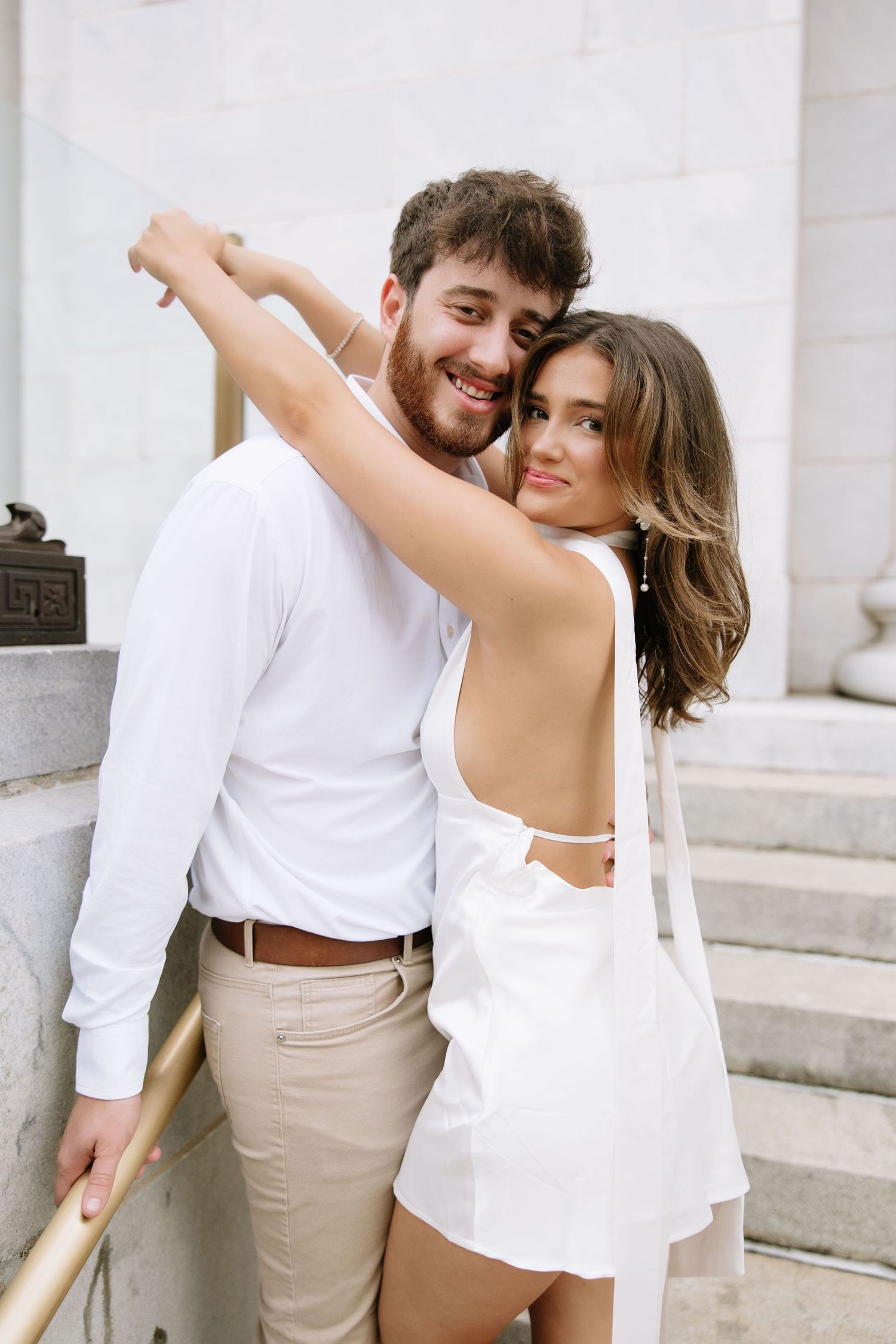 A couple in white outfits poses playfully on outdoor stone steps during what appears to be an engagement photoshoot.