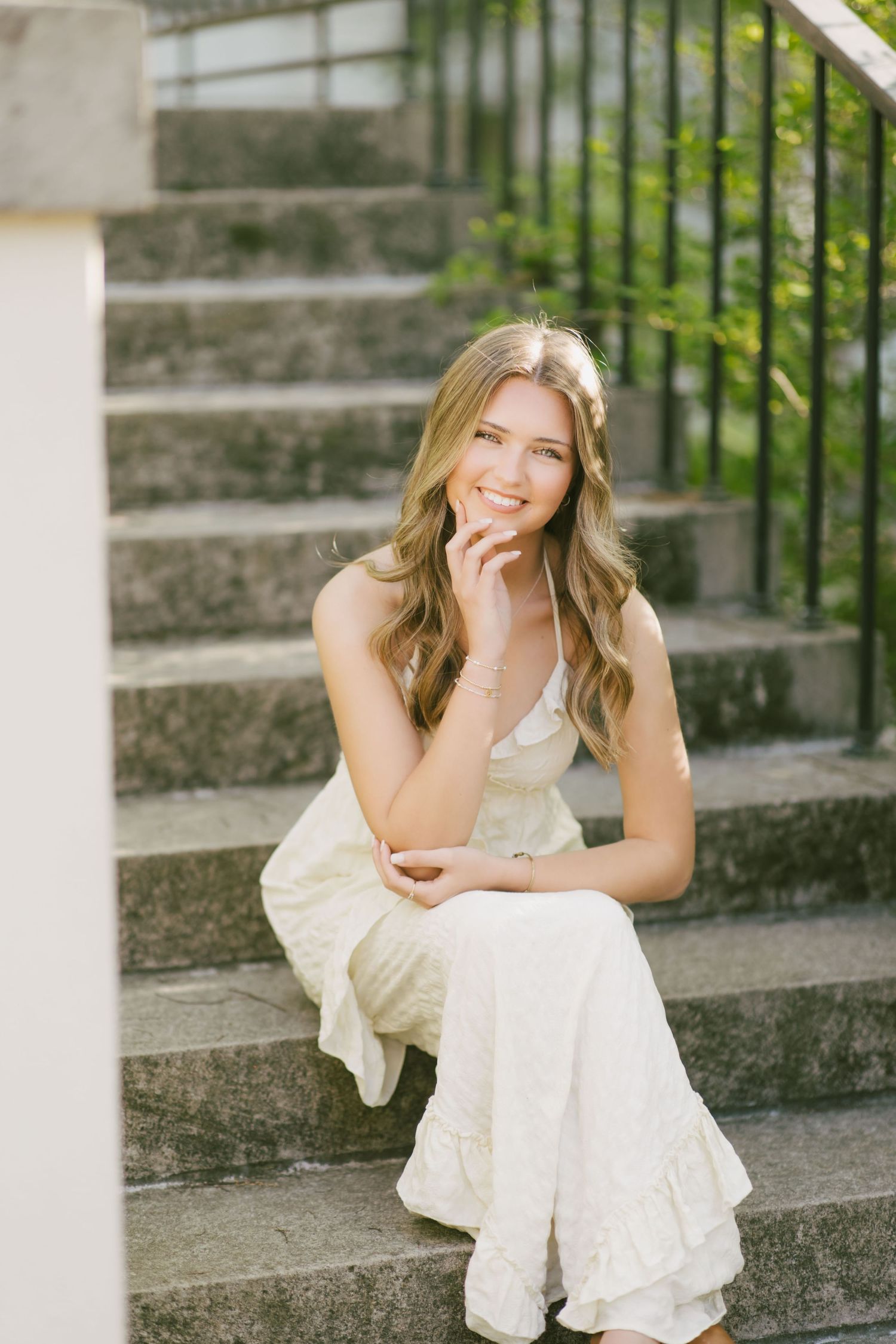 A young woman in a flowing cream-colored dress sits on stone steps outdoors with a natural, candid expression.