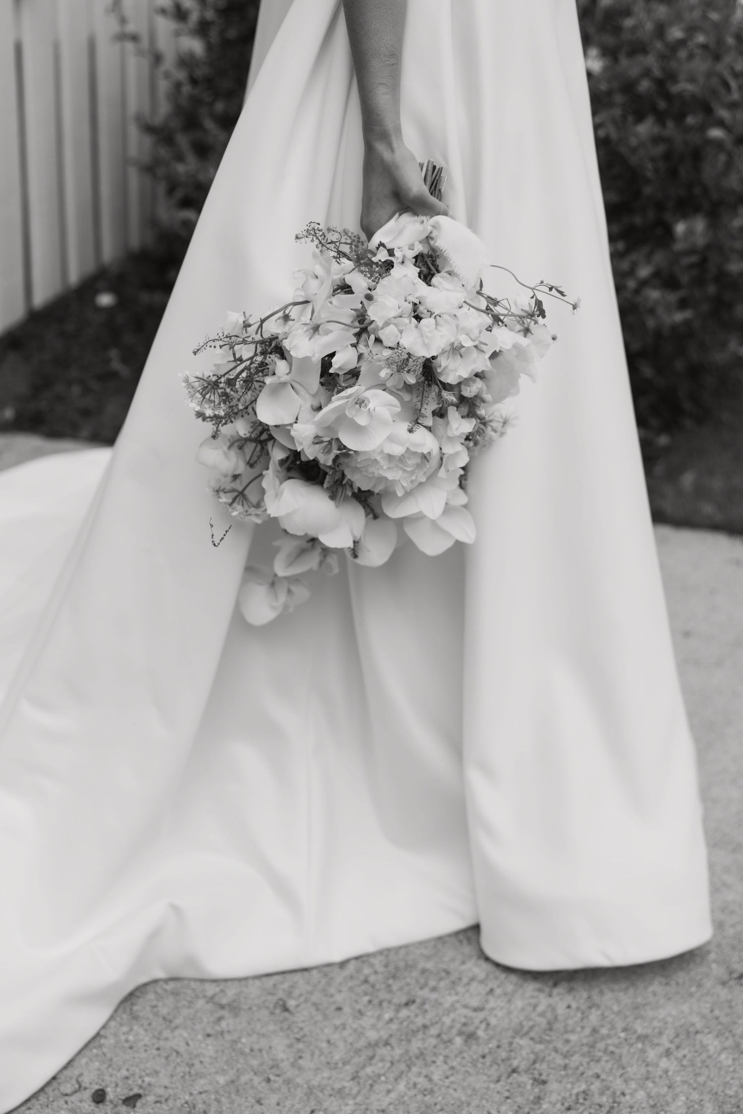 A black and white detail shot of an elegant white bridal bouquet against flowing wedding dress fabric.