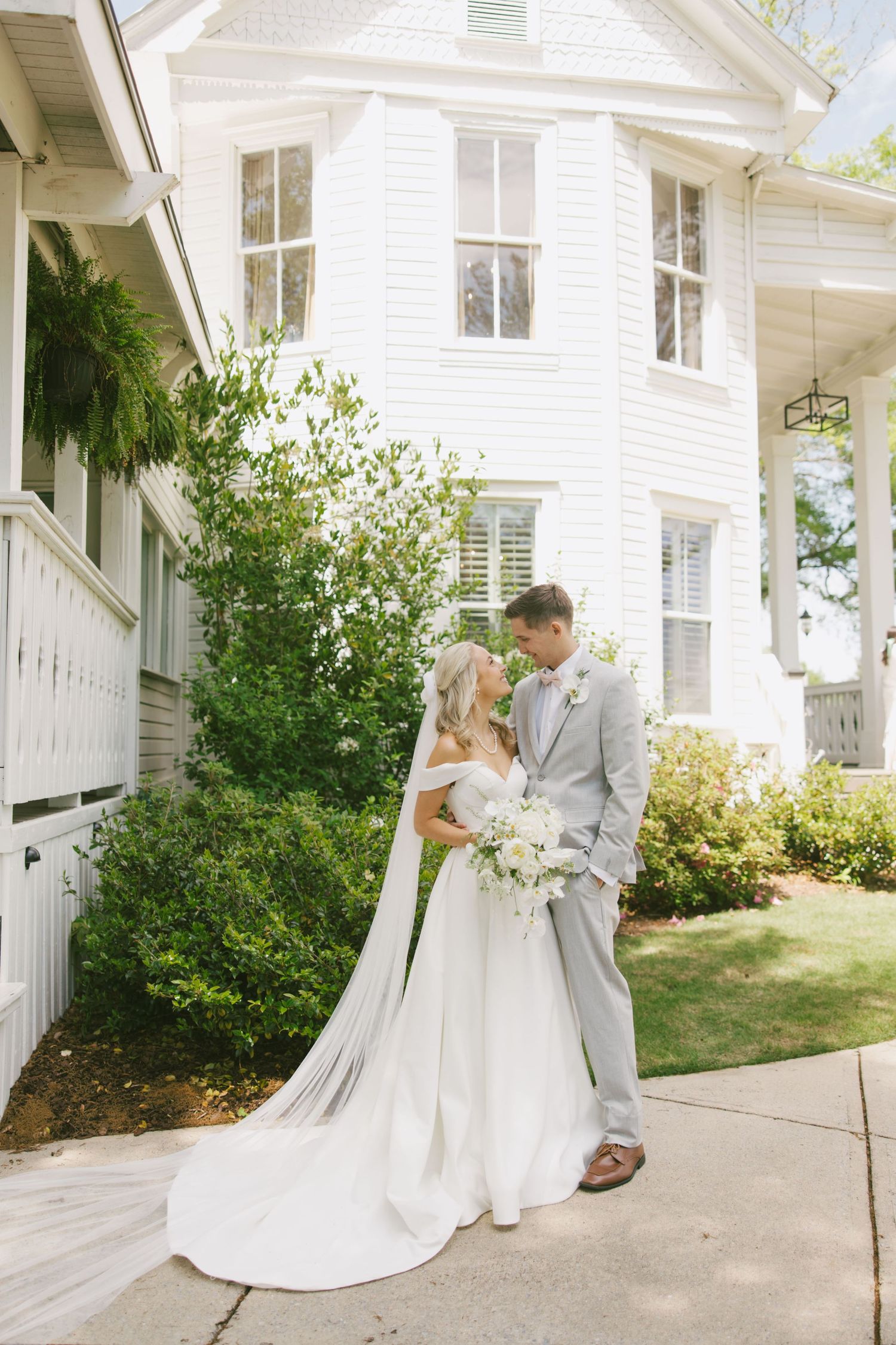 A bride and groom share a romantic moment outside a classic white colonial style house with flowing veil and florals.