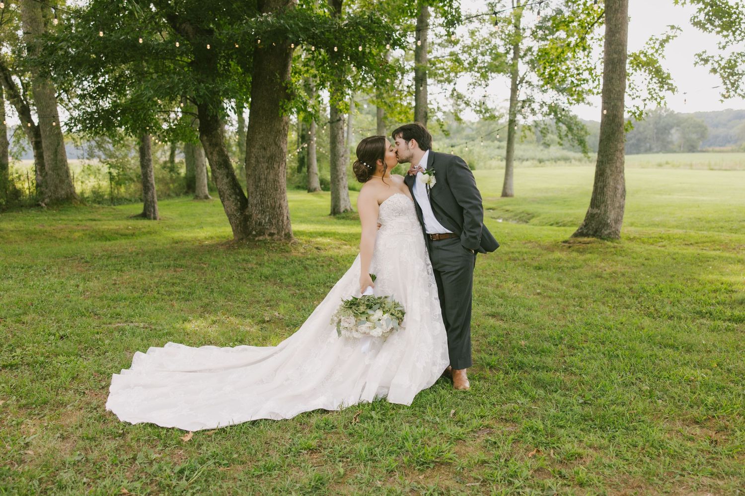 A bride and groom share an intimate moment together in a lush green field with tall trees during their outdoor wedding.