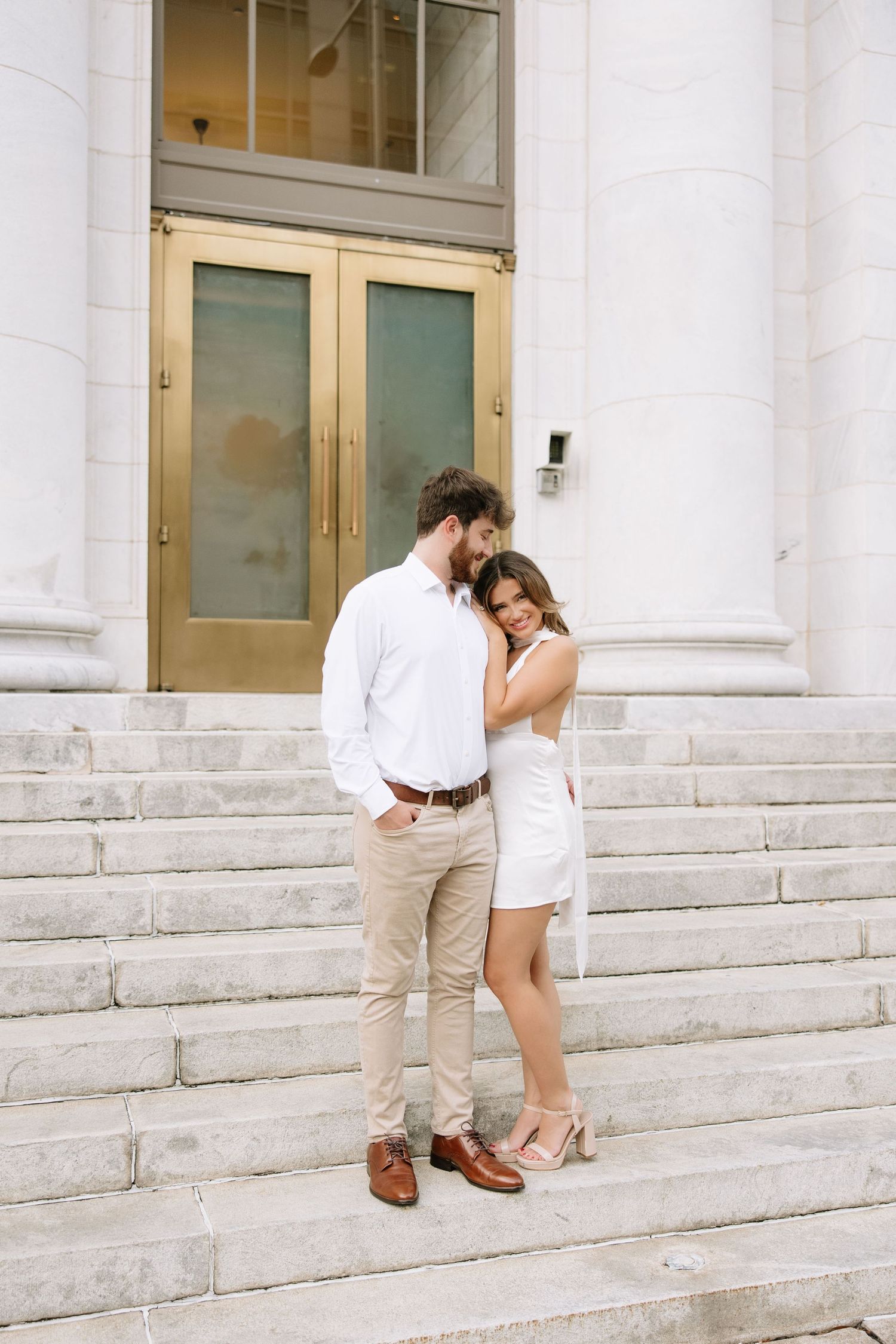 Couple in white and beige outfits embrace romantically on marble steps of a grand building entrance.