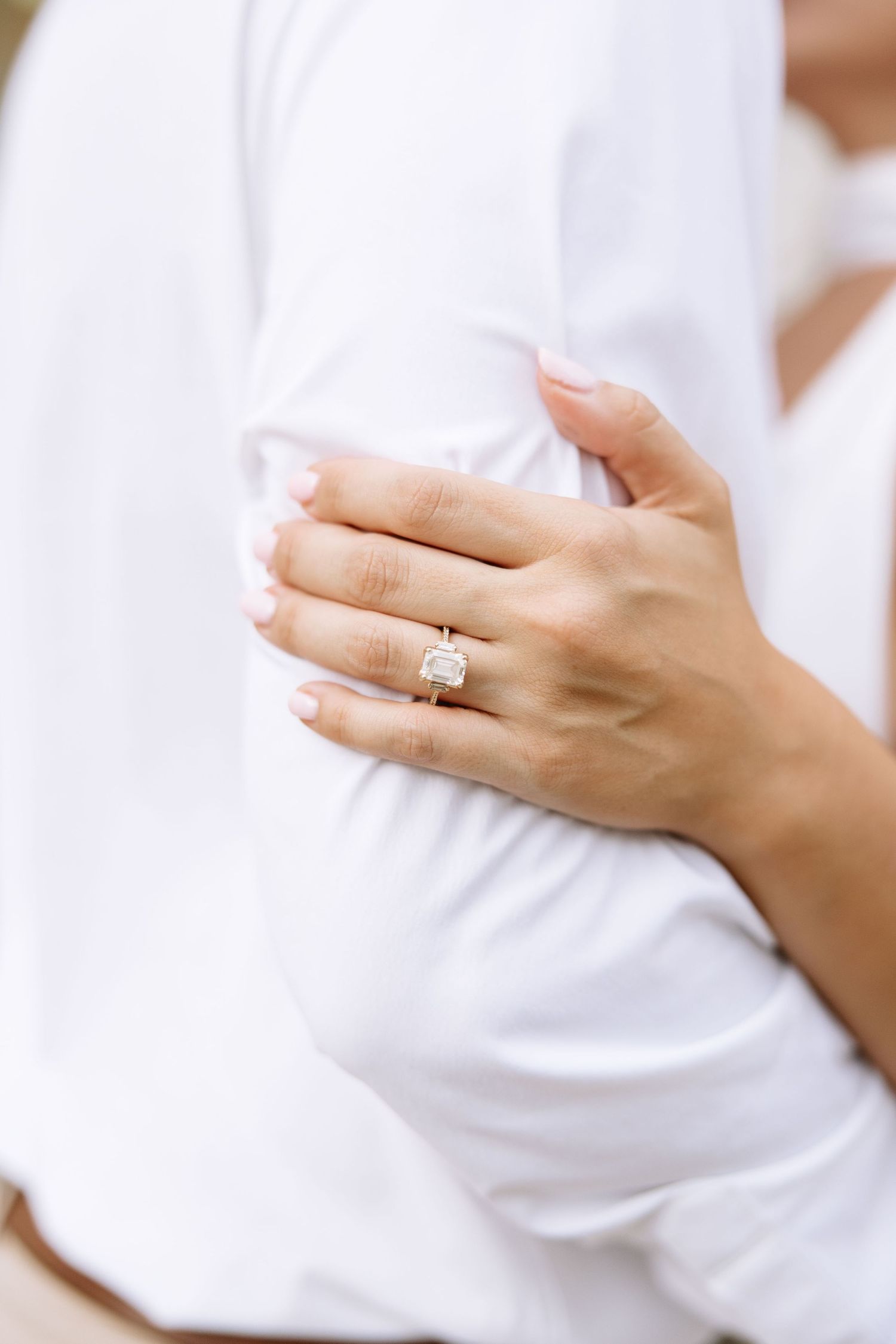 Close-up of feminine hands wearing an elegant engagement ring against white fabric background.