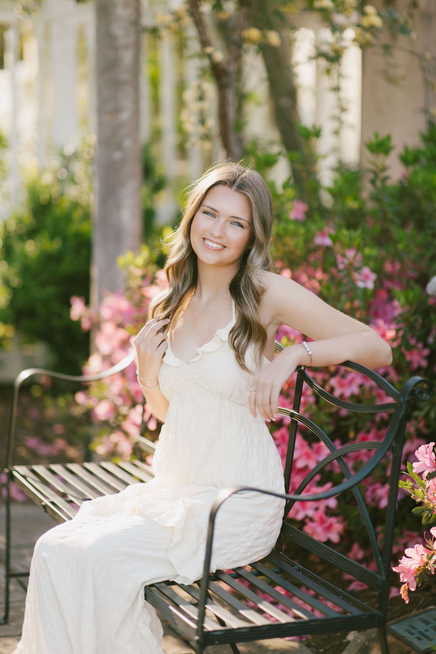 Someone in a flowing white dress sits on a decorative metal garden bench surrounded by blooming pink flowers and greenery.