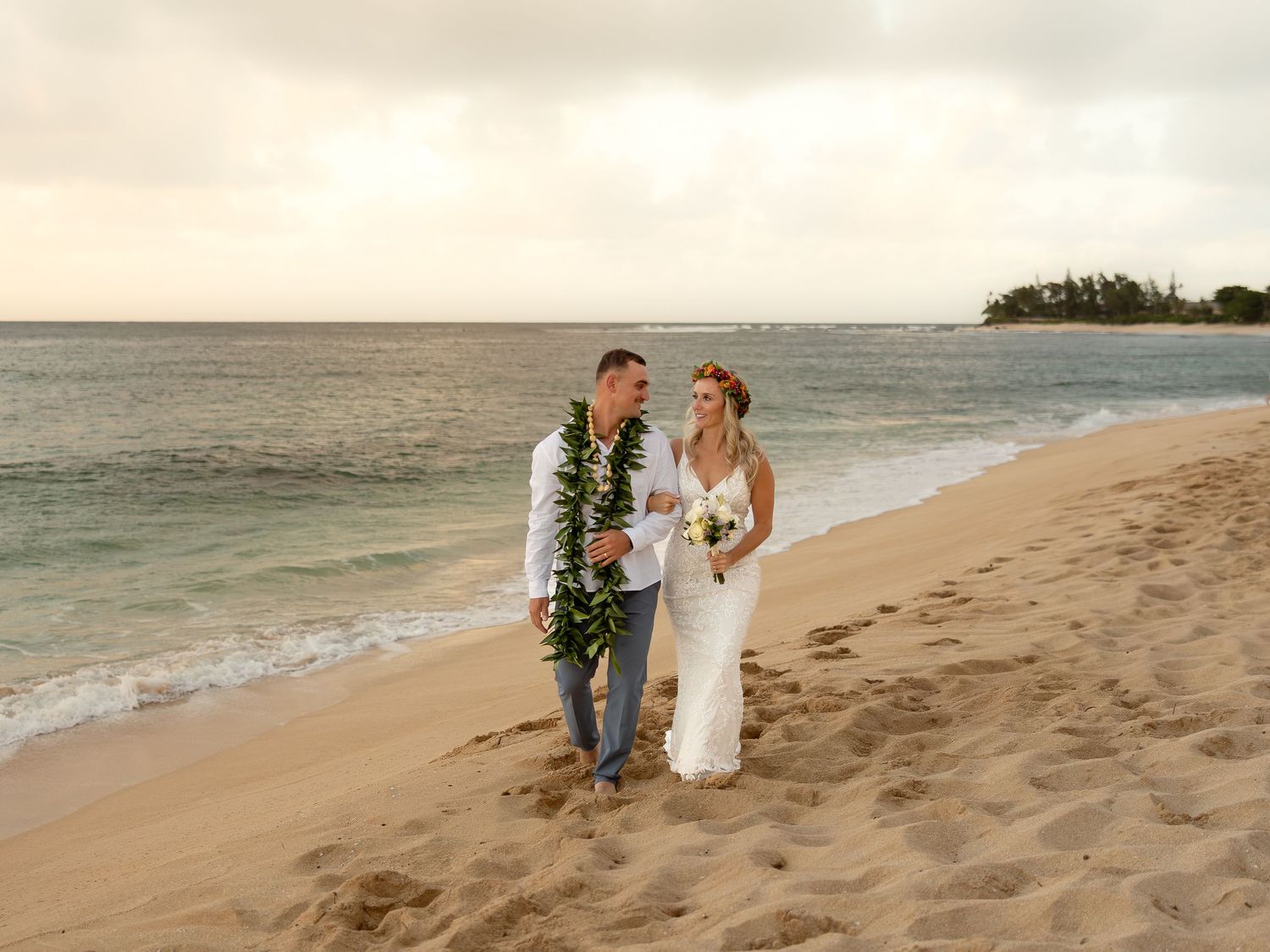 Newlywed couple walking on a sandy Hawaiian beach, groom wearing a lei, bride in a white gown, ocean backdrop.