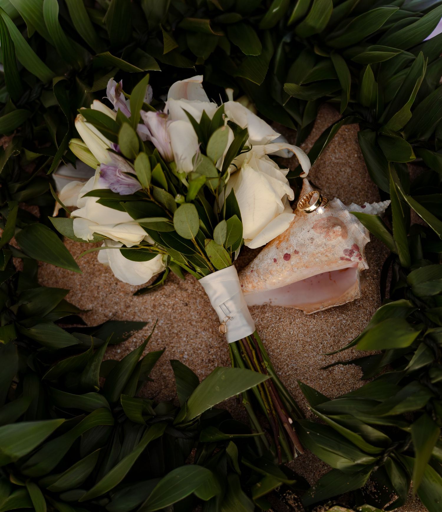 White calla lily and freesia wedding bouquet with seashell accent resting on sandy beach surrounded by tropical greenery.