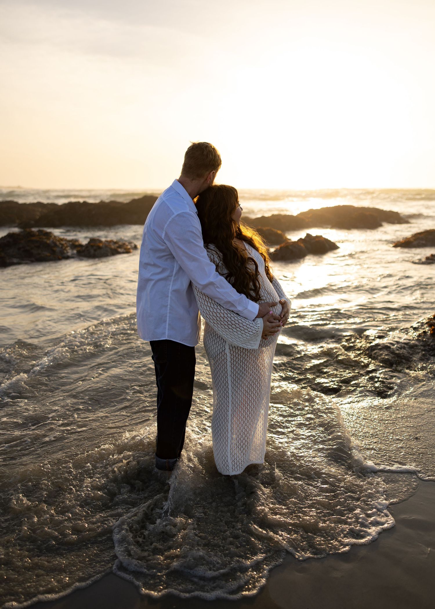 Couple embracing at sunset on a rocky beach, man in white shirt and woman in white dress, waves washing over their feet.