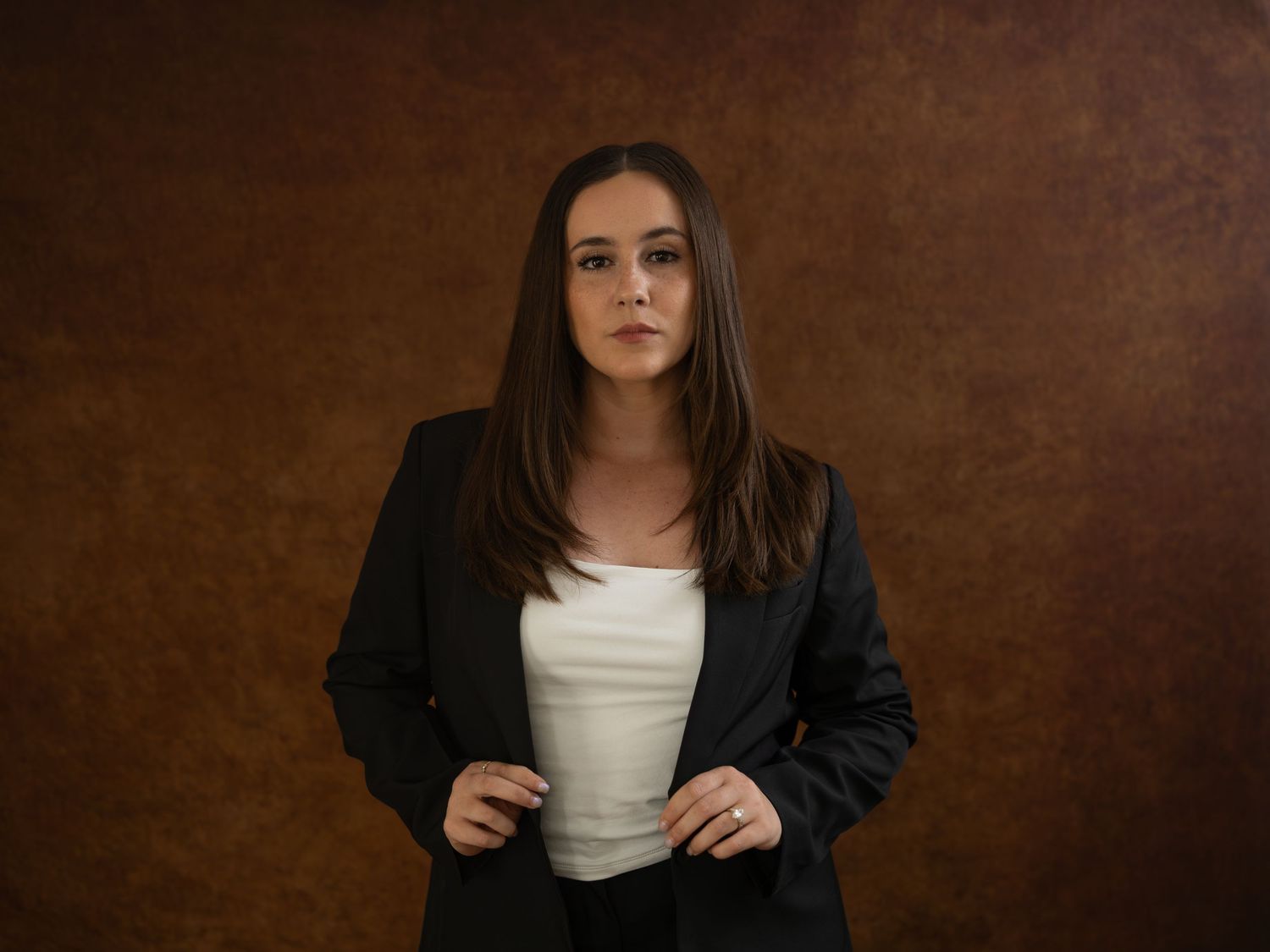 Professional woman in black blazer and white top posing against a brown textured studio background.