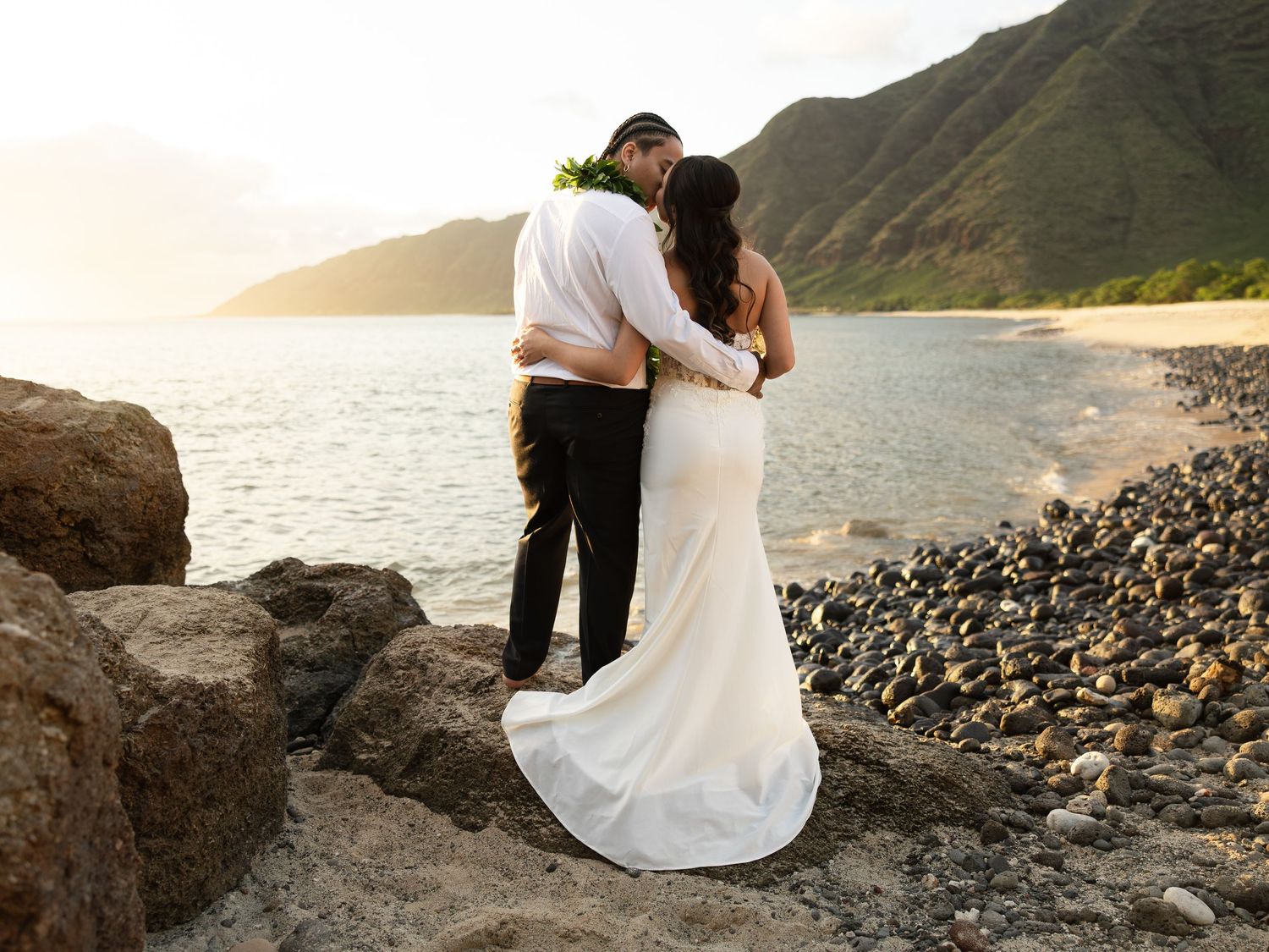 Newlywed couple embracing on rocky Hawaiian beach at sunset with mountains and ocean in background.