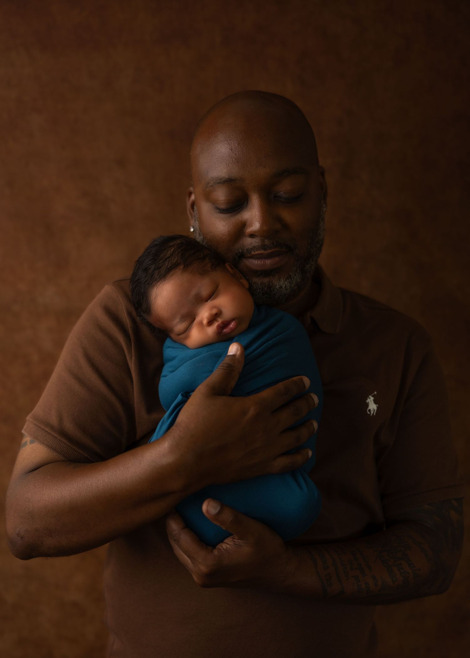 A bald father tenderly holds his swaddled newborn baby wrapped in teal against a warm brown studio backdrop.