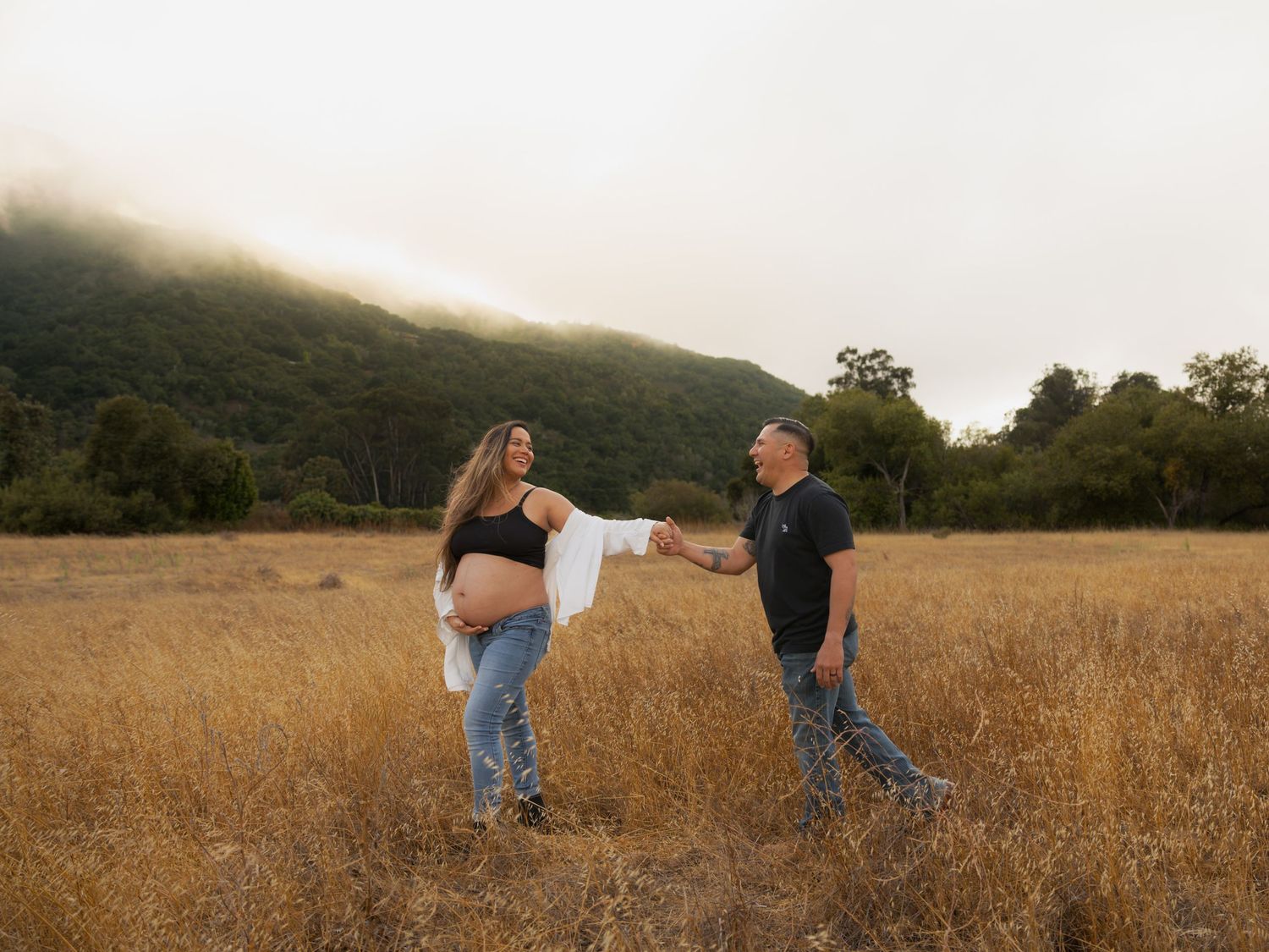 Pregnant couple holding hands in golden field with misty green hills, maternity photo shoot outdoors.