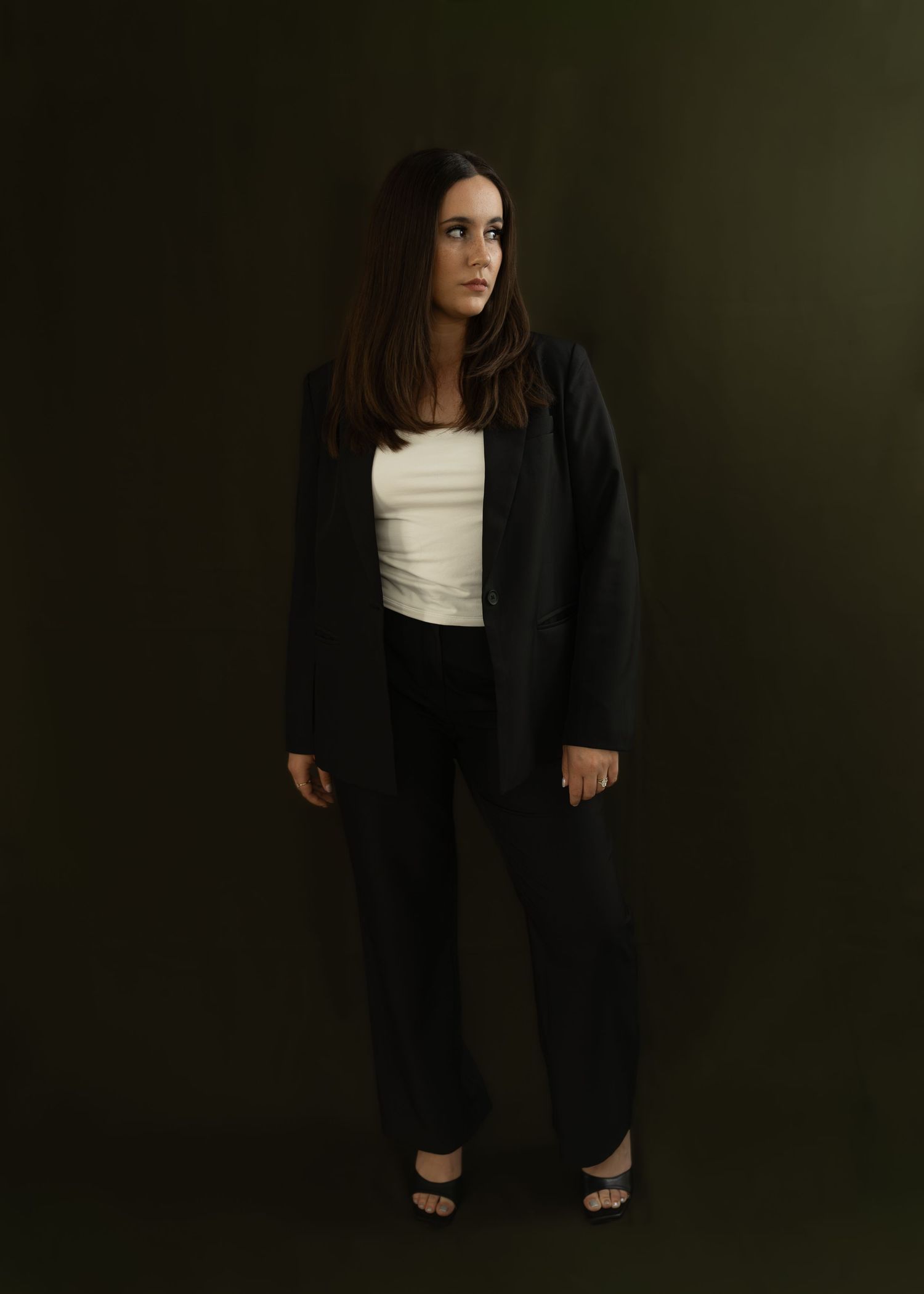 Woman in black blazer and trousers with white top stands against dark green background.