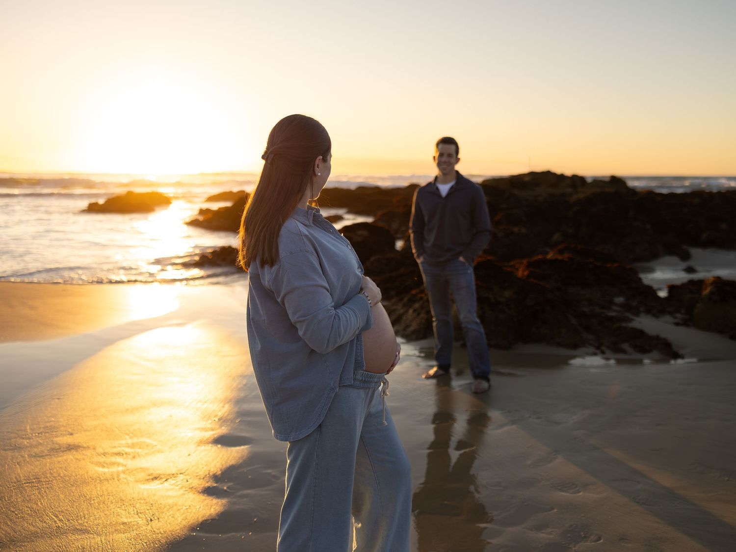 Pregnant woman and man share a tender moment on a beach at golden sunset, with rocky shore and ocean waves in background.