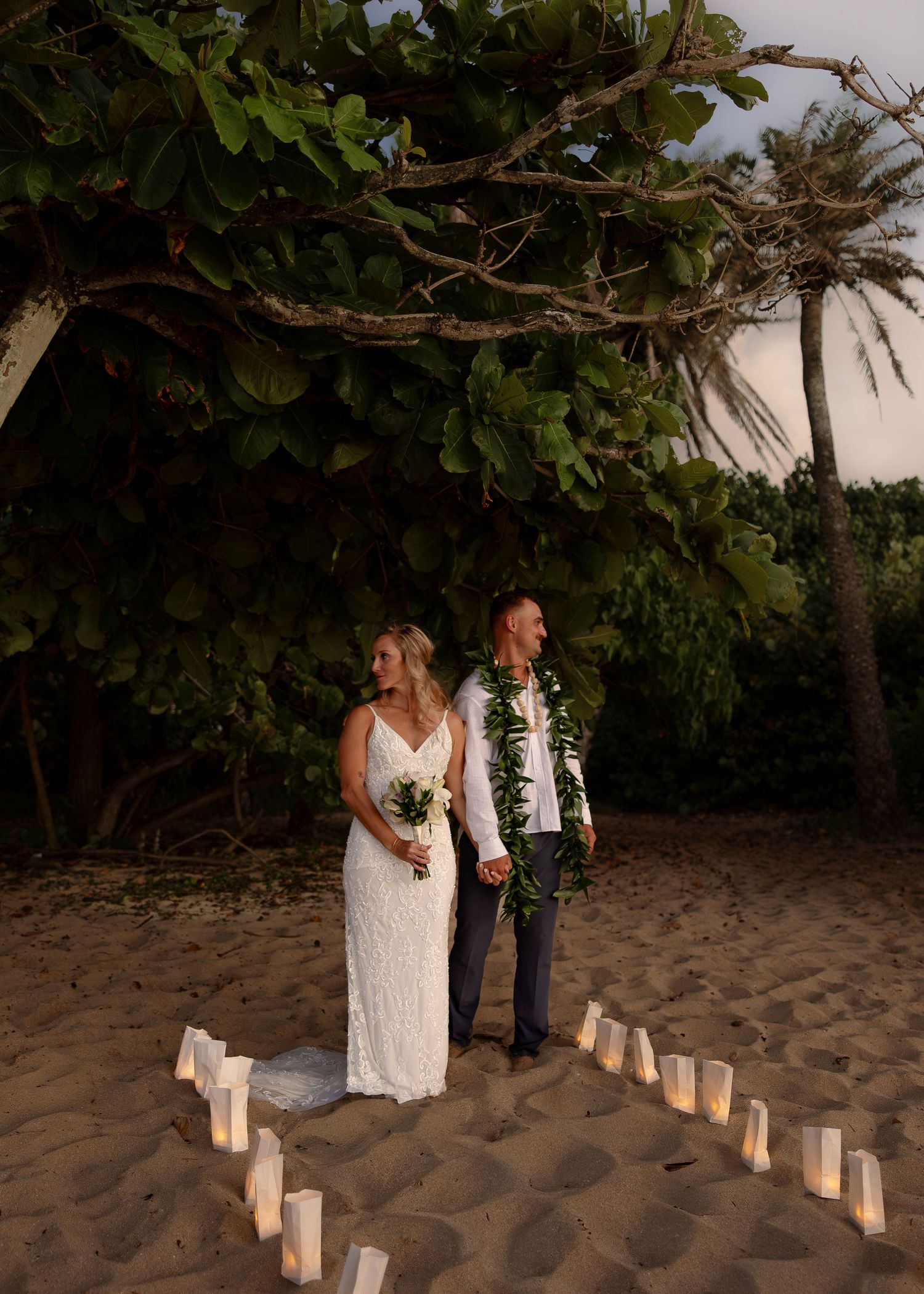 Bride and groom hold hands on sandy beach at dusk, surrounded by luminaries, beneath a lush tropical tree in Hawaii.