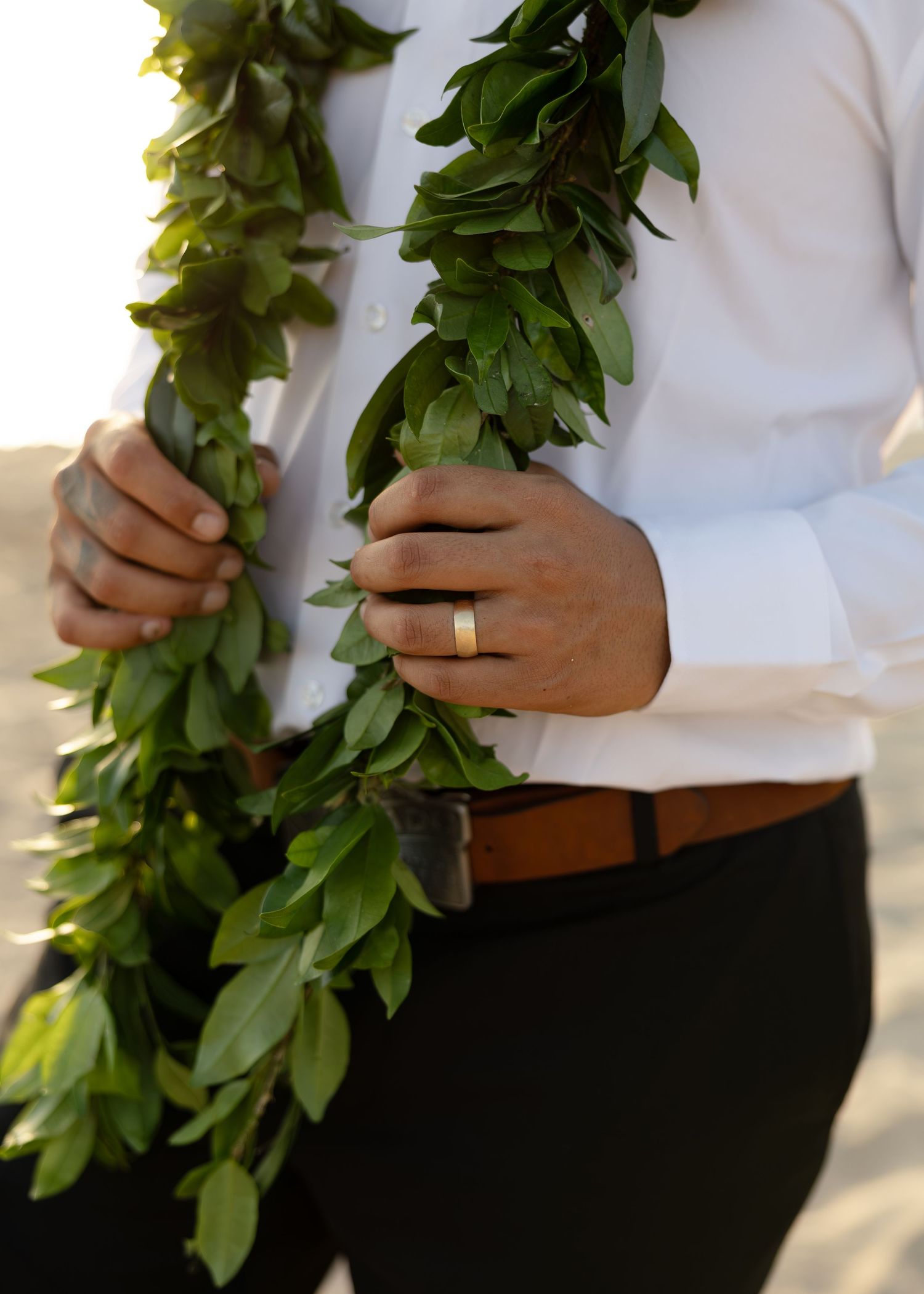 Groom in white shirt holding green leaf lei garland, wearing gold wedding band, on beach wedding day.