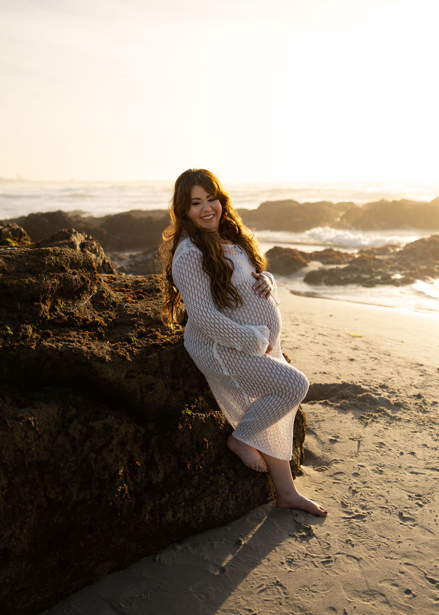 Pregnant woman in white lace dress sitting on beach rocks at golden hour sunset by the ocean.