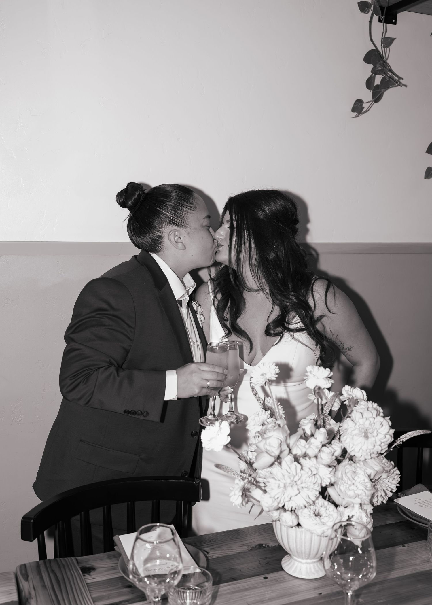 Black and white photo of a couple sharing a kiss at their wedding reception table with floral centerpiece.