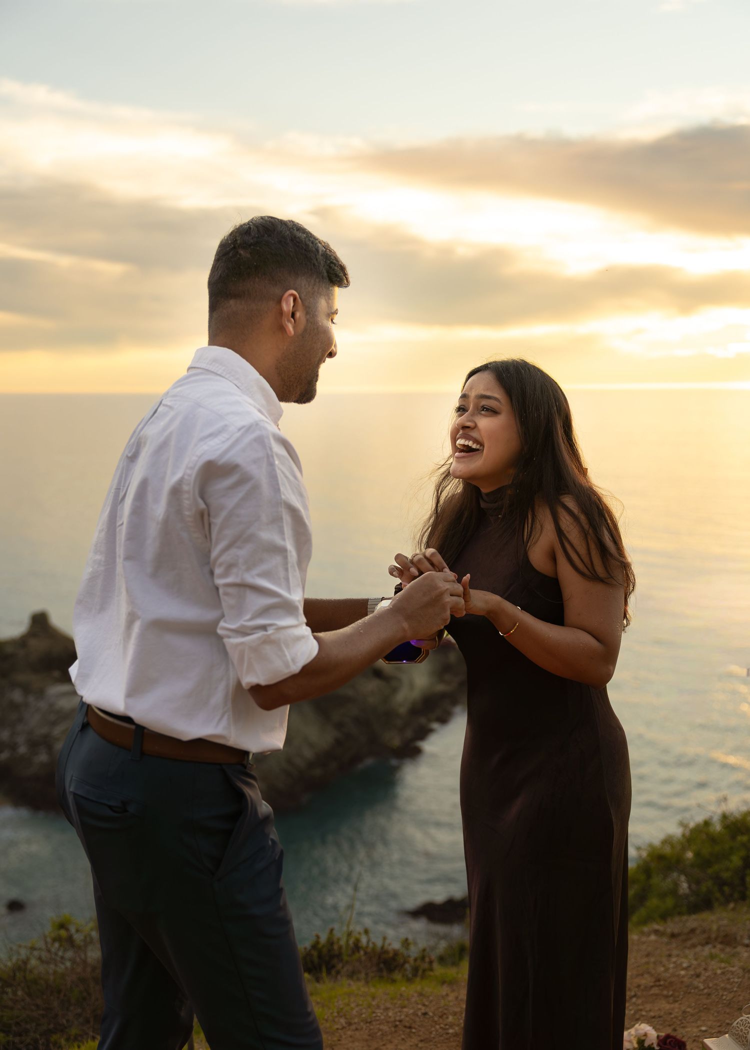 Couple sharing a romantic proposal moment at sunset on a coastal cliff with ocean views.