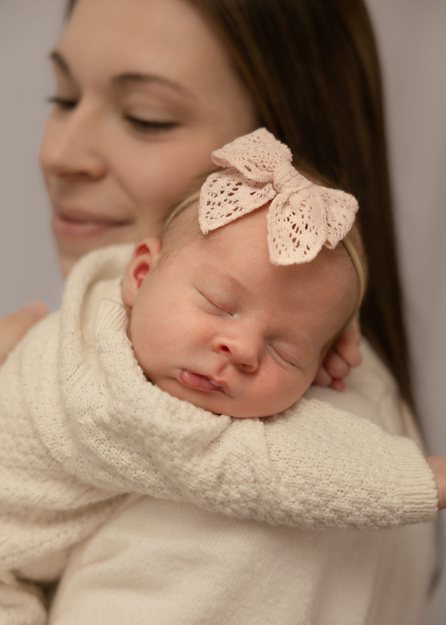 A mother tenderly holds her sleeping newborn baby wearing a delicate pink lace bow headband, wrapped in a cream knit blanket.