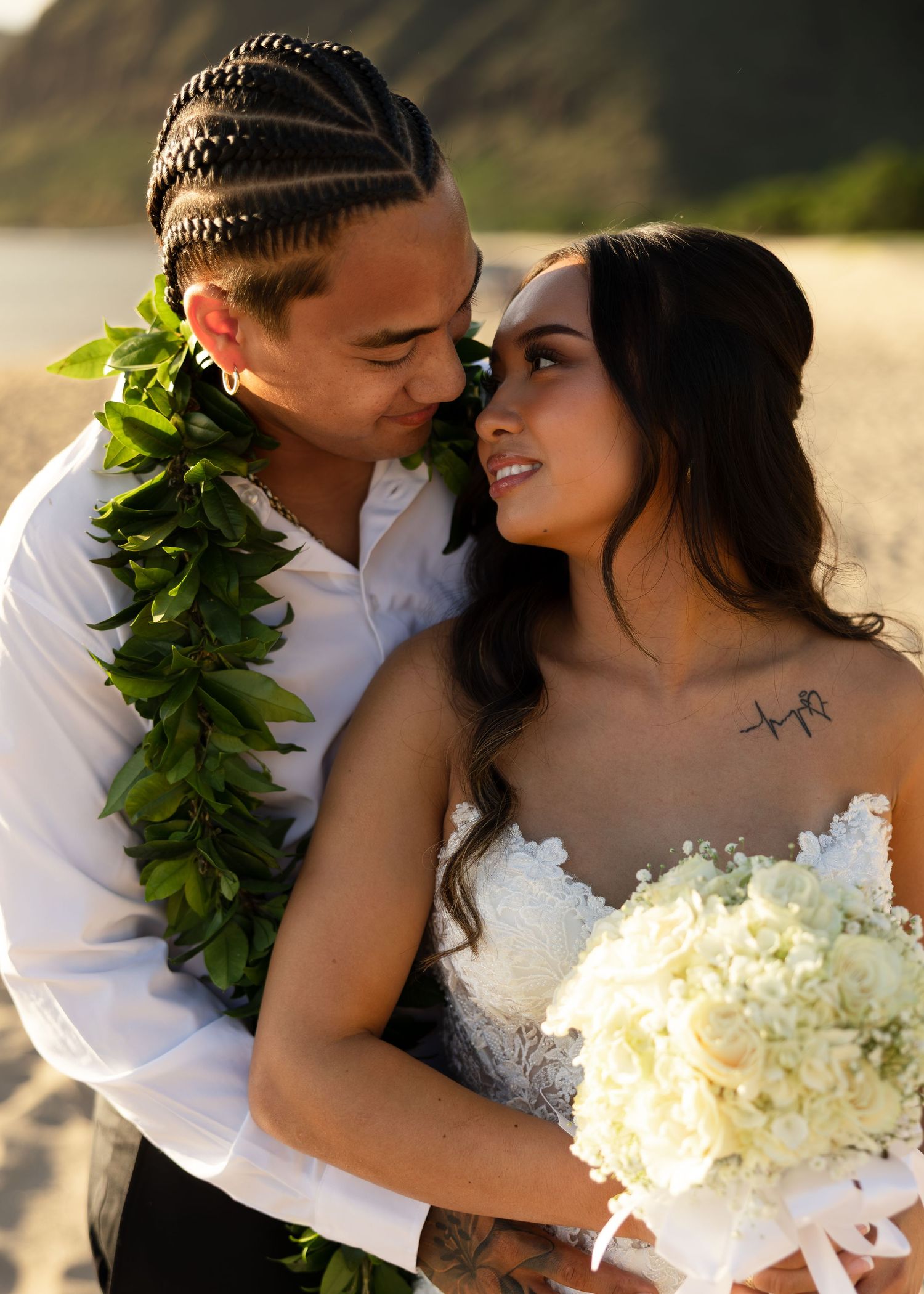 Couple in wedding attire embracing on beach, groom wearing green lei, bride holding white bouquet in Hawaii.