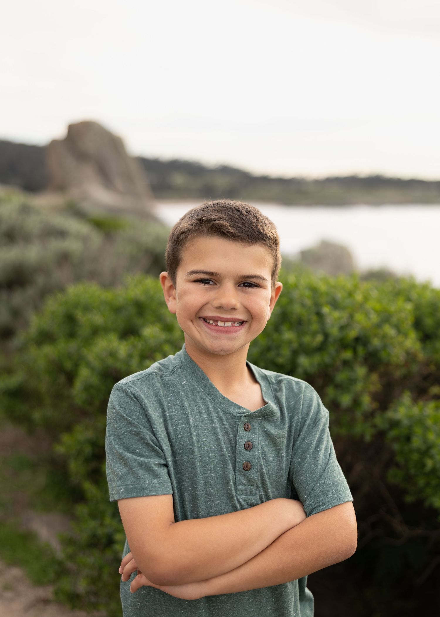 Smiling boy with crossed arms wearing a green henley shirt outdoors near coastal rocks and greenery.