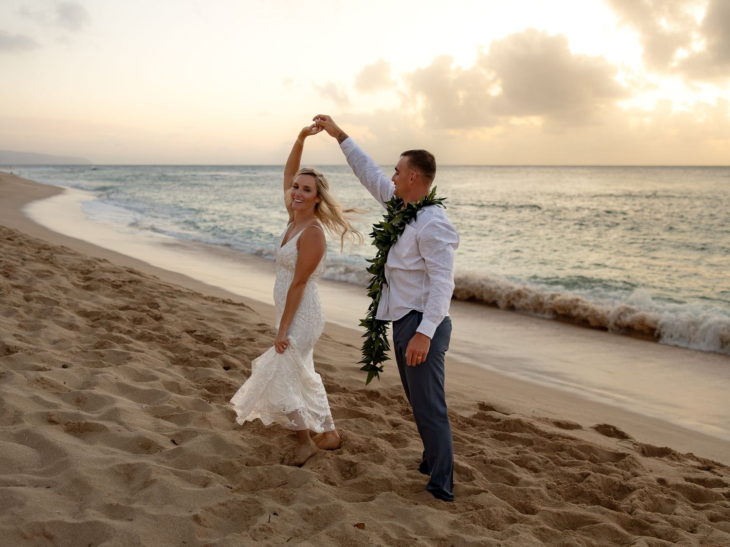 Newlywed couple dancing on a sandy beach at sunset, bride in white dress, groom with floral lei, ocean backdrop.