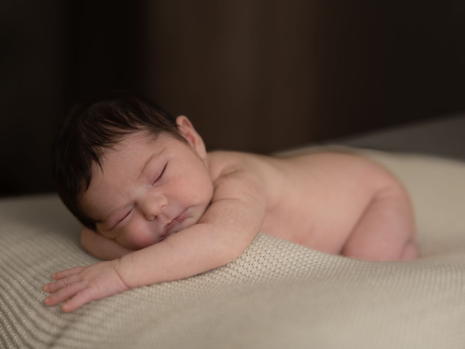 Peaceful newborn baby sleeping on a soft cream blanket, with dark hair and a gentle smile on their face.