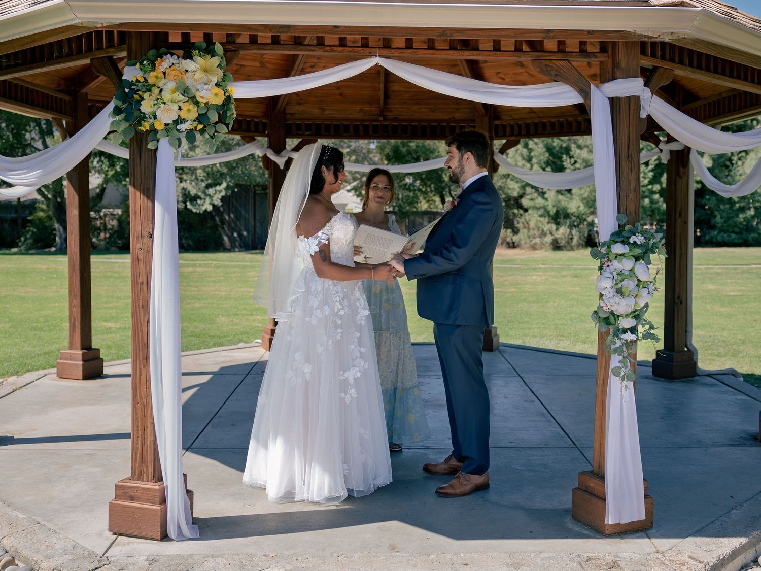Couple exchanging vows under a decorated gazebo with white draping and floral arrangements at an outdoor wedding ceremony.
