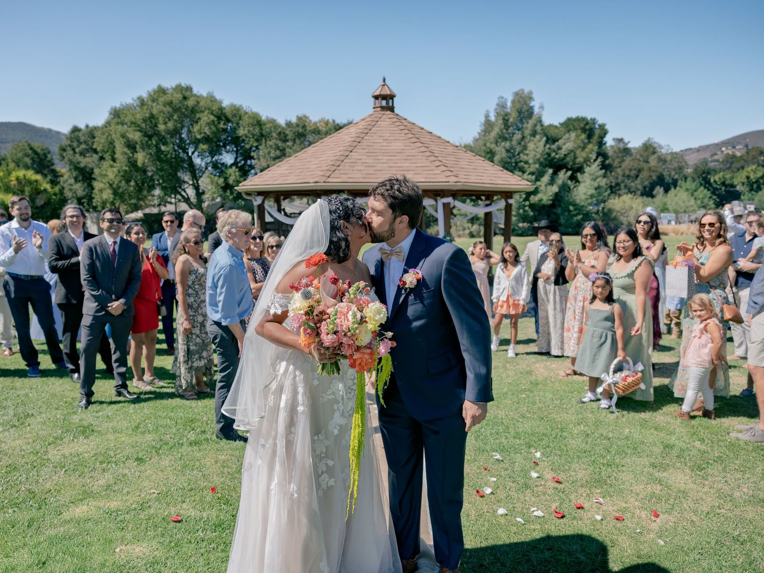 Bride and groom share a kiss outdoors at a garden wedding ceremony with guests lining the aisle near a gazebo.